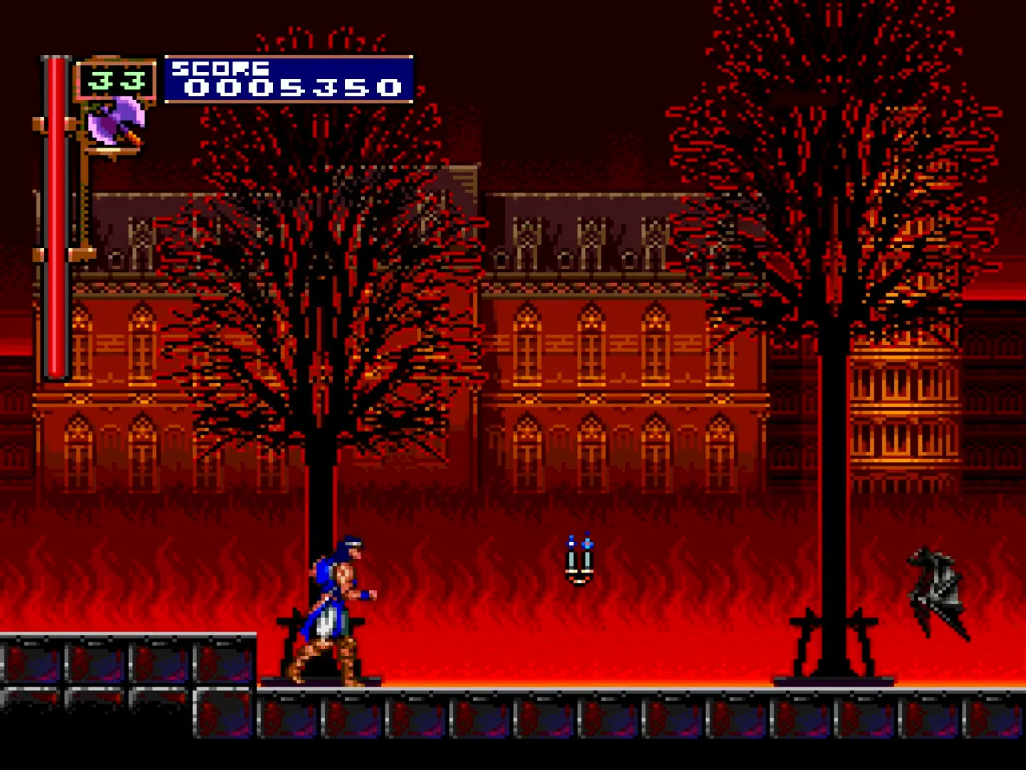 Richter Belmont walks forward through Stage 1's burning village. Two black skeletal trees flank gothic buildings under a red, flame-lit sky; a low fire-line crosses the cobbled foreground.